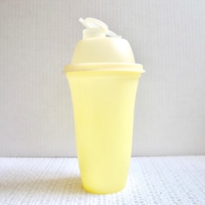 May include: A pale yellow plastic shaker bottle with a matching lid and a white flip-top closure. The bottle has a curved shape and is designed for mixing beverages or supplements. The shaker is approximately 8 inches tall.