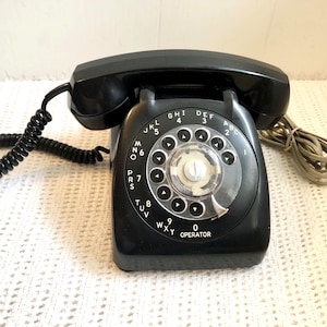 Może przedstawiać: Czarny telefon z tarczą obrotową z słuchawką, tarczą i zwiniętym kablem. Tarcza ma cyfry i litery, w tym "OPERATOR". Telefon ma klasyczny, zabytkowy design.