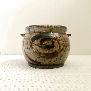 May include: A handmade ceramic pot with a speckled, earthy colour palette. The pot features a spiral design in shades of brown and tan. It has two small handles and sits on a textured surface, showcasing a rustic aesthetic.