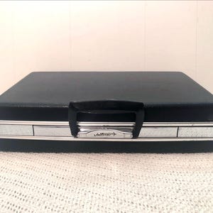 Puede incluir: Un maletín de máquina de escribir vintage negro con ribetes plateados y un asa negra. La palabra "Jelliner" está impresa en la parte delantera. El maletín es rectangular y está sobre una superficie blanca texturizada.
