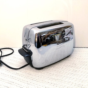 May include: A chrome General Electric toaster with two slots. The toaster has a black lever and a black power cord. The shiny surface reflects the surrounding environment. The toaster is on a white surface.