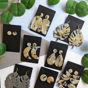 May include: Assortment of handmade polymer clay earrings in various shapes and colors, including gray, black, and white with gold accents. The earrings are displayed on black cards with gold hooks.