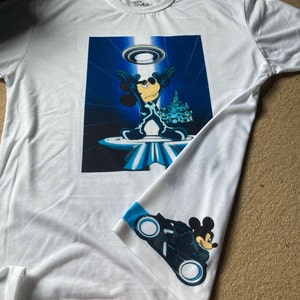 May include: White t-shirt featuring a blue and white graphic of Mickey Mouse in a futuristic setting. The shirt also has a blue and white graphic of Mickey Mouse riding a futuristic motorbike on the sleeve.