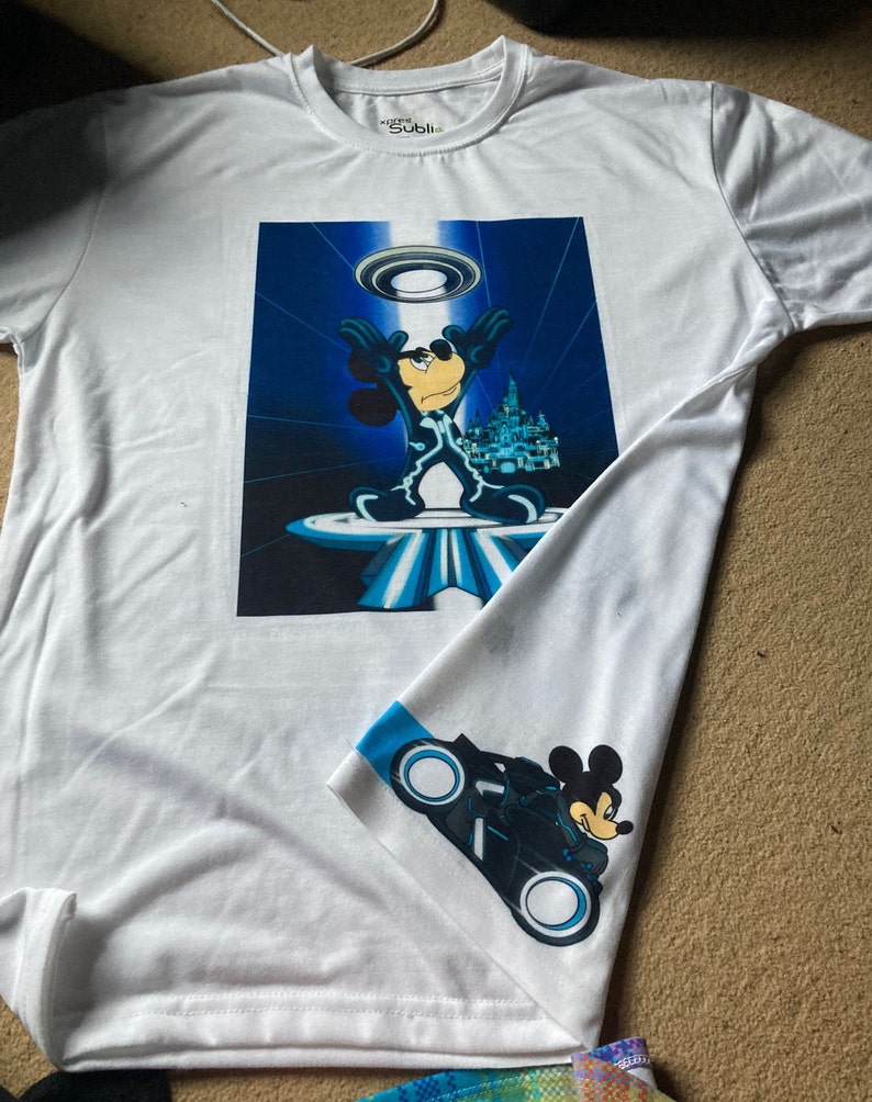 May include: White t-shirt featuring a blue and white graphic of Mickey Mouse in a futuristic setting. The shirt also has a blue and white graphic of Mickey Mouse riding a futuristic motorbike on the sleeve.