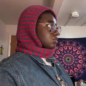 Könnte beinhalten: Eine gestrickte Kapuze in rot-grauen Streifen. Die Kapuze bedeckt Kopf und Hals und besteht aus einem weichen, strukturierten Garn. Die Kapuze ist Teil einer Winterbekleidungskollektion.