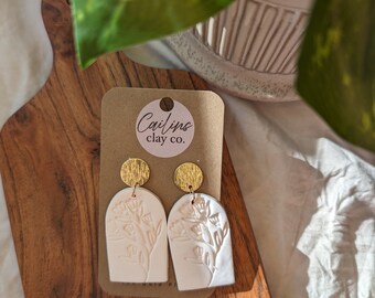 May include: A pair of off-white clay earrings with floral designs, attached to gold-colored circular studs. The earrings are displayed on a brown wooden cutting board, with a tag that reads "Cailins clay co."