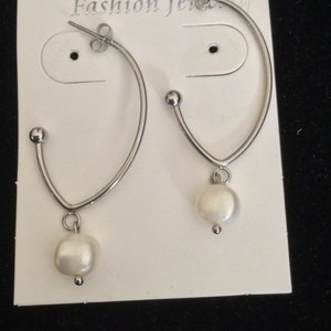 Puede incluir: Un par de pendientes de aro de plata con una sola perla blanca colgando de cada aro. Los pendientes se muestran en una tarjeta blanca con el texto "Fashion Jewelry".