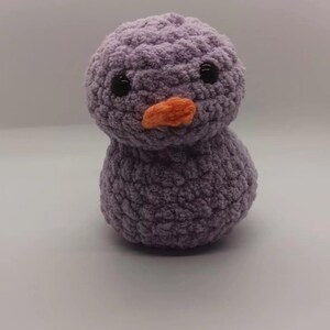 May include: A handmade, lavender-colored crochet duckling with black eyes and an orange beak. The plush toy has a rounded body and head, and is made of a soft, textured yarn. This adorable duckling is a perfect gift for children.