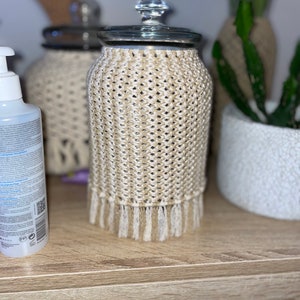 May include: A glass jar with a crocheted cover in beige and white. The jar has a glass lid and a crocheted fringe.