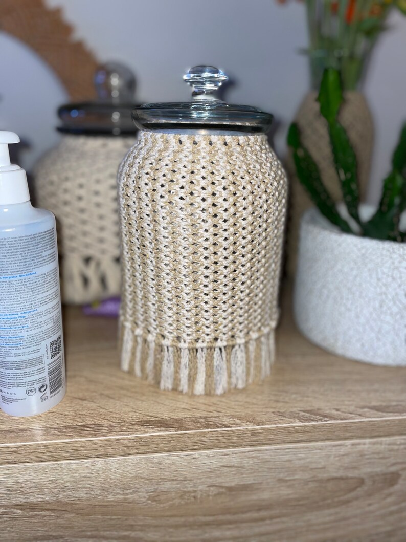 May include: A glass jar with a crocheted cover in beige and white. The jar has a glass lid and a crocheted fringe.