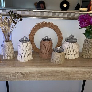 May include: Five glass jars with crocheted covers in white and beige with fringe. The jars are arranged on a wooden table with a wooden tray in the background.