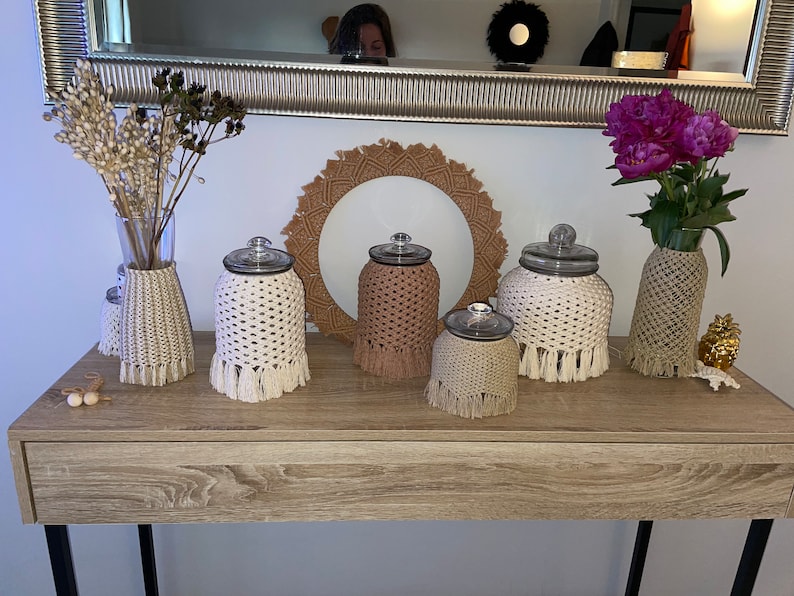 May include: Five glass jars with crocheted covers in white and beige with fringe. The jars are arranged on a wooden table with a wooden tray in the background.