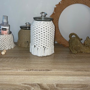 May include: Three glass jars with white crocheted covers and fringe. The jars are on a wooden surface with a brown woven circle and a gold decorative object.