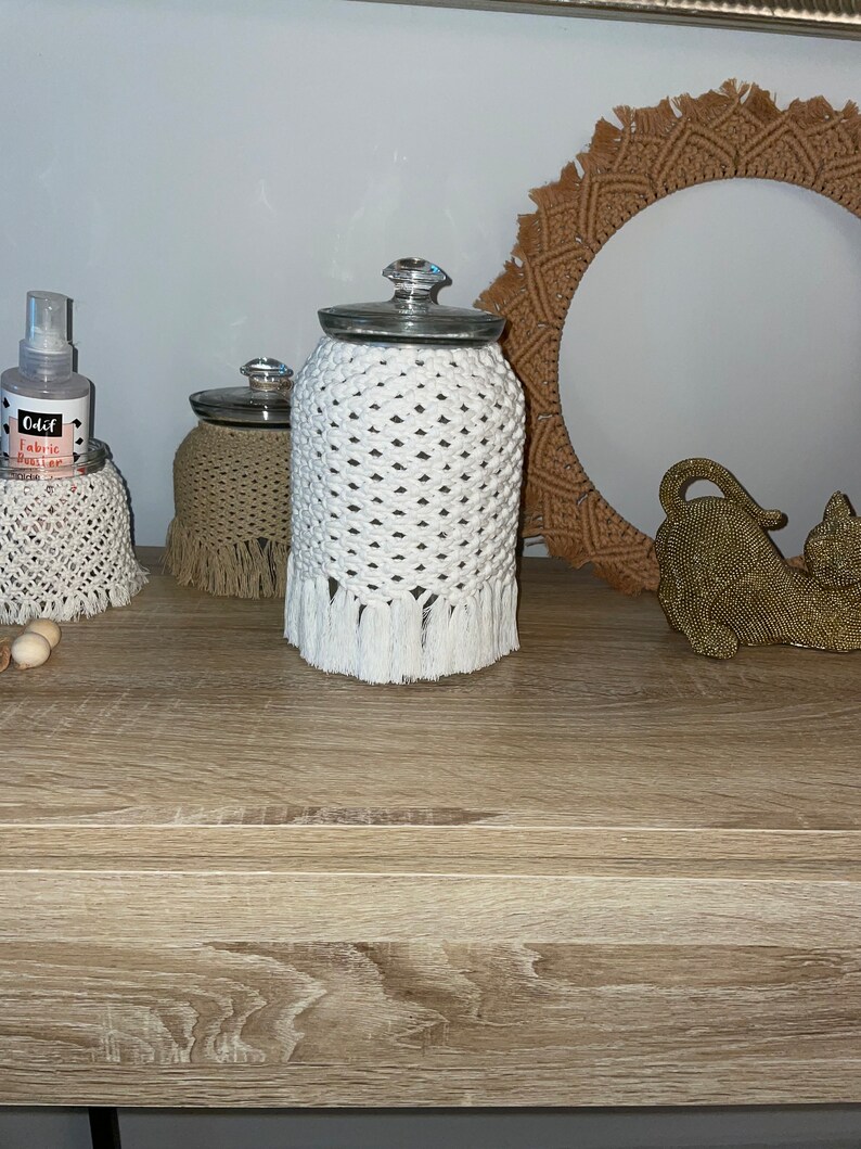 May include: Three glass jars with white crocheted covers and fringe. The jars are on a wooden surface with a brown woven circle and a gold decorative object.