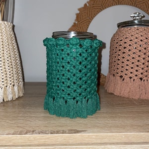 May include: A green crocheted jar cozy with a fringe border and a row of decorative knots around the top. The cozy is on a wooden surface.
