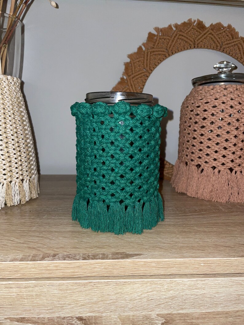 May include: A green crocheted jar cozy with a fringe border and a row of decorative knots around the top. The cozy is on a wooden surface.