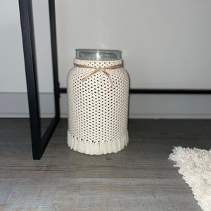 May include: A white crocheted jar cover with a brown ribbon tied around the top. The jar is sitting on a wooden surface.