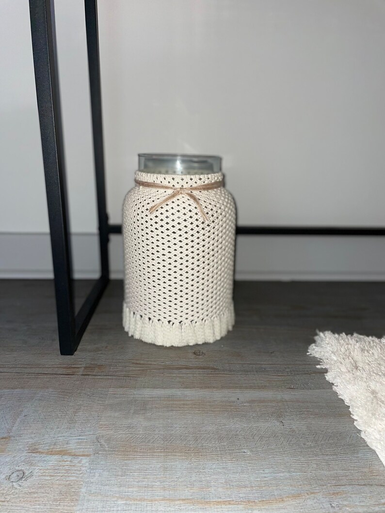 May include: A white crocheted jar cover with a brown ribbon tied around the top. The jar is sitting on a wooden surface.