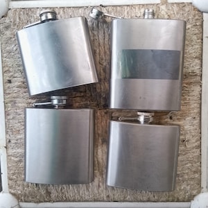 Op de afbeelding: Vier zilveren metalen hipflasks met een schroefdop. De flasks zijn in een vierkant patroon op een houten oppervlak gerangschikt.