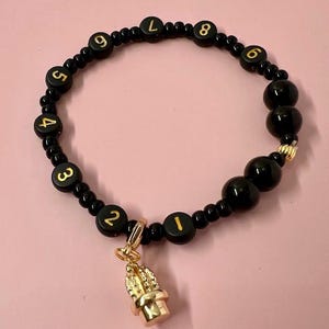 May include: Black beaded bracelet with gold numbers and a gold charm. The bracelet features black beads with gold numbers from 1 to 9. A gold charm hangs from the bracelet. The bracelet is on a pink background.
