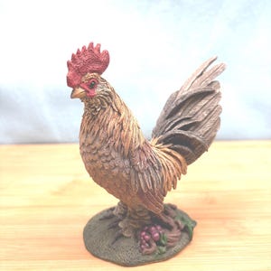 May include: A detailed figurine of a rooster, featuring a red comb and wattle, with brown and tan feathers. The rooster stands on a green base with grapes. The sculpture is set against a light background and wooden surface.