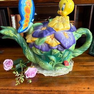 May include: A ceramic teapot featuring a green vine-textured body with a small red ladybug, a lid adorned with yellow and purple flowers, a yellow Tweety Bird figure, and a large blue and orange butterfly attached to the side of the spout.