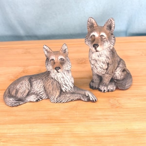 May include: Two detailed wolf figurines. One is lying down, the other is sitting upright. Both are painted in shades of gray, brown, and white, with realistic features. The figurines are placed on a wooden surface.