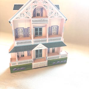 May include: A miniature pink Victorian house with intricate architectural details. Features include a gray roof, blue shutters, and decorative trim. The house has multiple balconies and a small front porch with a lavender garden.