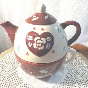 May include: A ceramic tea for one set featuring a cream-colored teapot with a brown lid, handle, and spout. The teapot displays a central brown heart with a white rose design, accented by small scattered heart and leaf motifs. A matching cup is included.