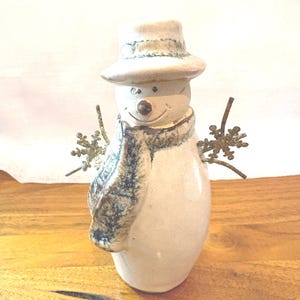 May include: A ceramic snowman figurine with a smooth off-white finish, wearing a matching hat and a textured scarf with blue and brown accents. The snowman has a small brown nose, two dark eyes, and metallic gold snowflake-shaped arms.