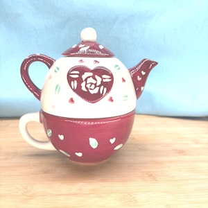 May include: A ceramic tea-for-one set. The teapot is white with a burgundy heart-shaped design containing a rose. The cup is burgundy with white and green accents. The lid has a white knob and burgundy accents. The set is on a wooden surface.