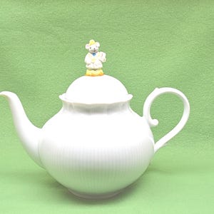 May include: White porcelain teapot with a lid and a small figurine on top. The teapot has a curved spout and handle. The figurine is a small bear wearing a hat and holding a book. The teapot is on a green background.