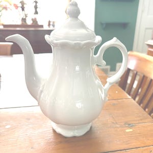 May include: A white ceramic teapot with a lid, spout, and handle. The teapot has a decorative, scalloped edge around the lid and base. The handle is curved, and the spout is long and slender. The teapot is sitting on a wooden table.
