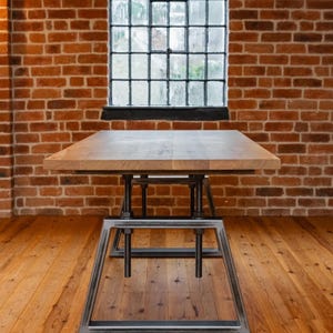 Op de afbeelding: Een rechthoekige houten tafel met een metalen onderstel. De tafel staat tegen een bakstenen muur met een raam. De afmetingen van de tafel zijn ongeveer 152 cm lang, 76 cm breed en 76 cm hoog.