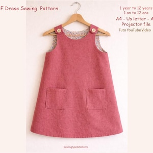 May include: A dusty rose dress pattern for ages 1 to 12 years, displayed on a wooden hanger. The dress features two front pockets and button details on the straps. The image includes the text "PDF Dress Sewing Pattern" and "Sewing Spells Patterns".