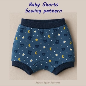 Peut inclure: Short de bébé bleu avec un motif de points blancs, d'étoiles jaunes et d'ours noirs. Le short a une taille élastique bleu marine et une bordure bleu marine autour des jambes. Le texte "Baby Shorts Sewing pattern" est en haut de l'image.