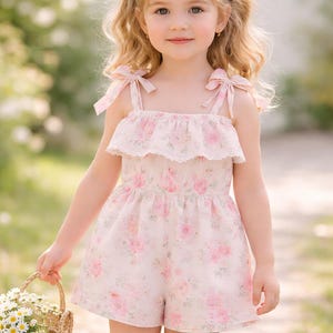 Peut inclure: Une jeune enfant porte une barboteuse rose clair à motifs floraux et garniture en dentelle. La barboteuse a des bretelles nouées et un col à volants. L'enfant tient un petit panier en osier rempli de fleurs blanches.