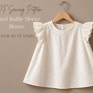 Peut inclure: Un chemisier blanc en broderie anglaise avec des manches à volants évasées suspendu à un cintre en bois. Le texte sur l'image indique : PDF Sewing Pattern, Flared Ruffle Sleeve Blouse, 1 year to 12 years. Il présente un col rond et un motif floral.