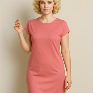 Op de afbeelding: Een koraalkleurige t-shirtjurk met korte mouwen en een ronde hals. De jurk is gemaakt van een zachte, effen stof en heeft een eenvoudig, klassiek ontwerp. Het model draagt de jurk.