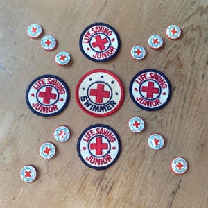 Puede incluir: Un conjunto de parches bordados y pines redondos con cruces rojas. Los parches dicen "Life Saving Junior" y "Swimmer". Los pines dicen "Beginner Swimmer" e "Intermediate Swimmer".