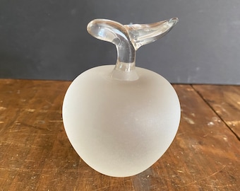 May include: A frosted glass apple shaped bottle with a clear glass stem. The bottle is a light white color and has a smooth, rounded shape.