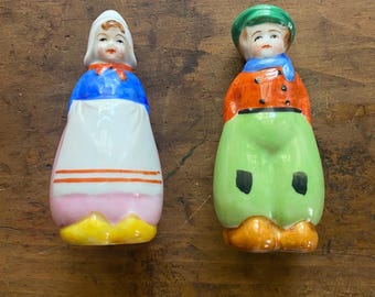 Könnte beinhalten: Zwei Keramikfiguren, die holländische Figuren darstellen. Eine ist eine Frau mit weißer Haube, blauem Oberteil und weißer Schürze. Die andere ist ein Mann mit grünem Hut, orangefarbener Jacke und grüner Hose. Beide haben bemalte Gesichter und stehen aufrecht.