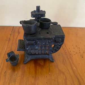 May include: Black cast iron toy stove with a pot, pan, and a small bucket. The stove has the words "AAA Queen" on the front.