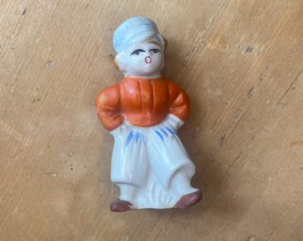 Puede incluir: Pequeña figura vintage de una persona con vestimenta tradicional. La figura presenta un tocado azul claro, una chaqueta naranja y pantalones blancos con detalles azules. La figura tiene zapatos marrones y posa con las manos en las caderas.