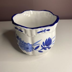 May include: White ceramic planter with a scalloped rim and blue floral design. The planter has a blue rim and blue floral accents. A decorative container for plants or flowers.