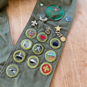 May include: A green Boy Scout sash with various merit badges and patches, including a fingerprint, a hammer and nails, a first aid cross, and a camping scene. The sash also features a gold star and a patch with the words "Boy Scouts of America".