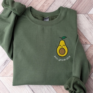 Könnte beinhalten: Ein grünes Sweatshirt mit einem niedlichen gestickten Avocado-Motiv und dem Text "Avo great day".