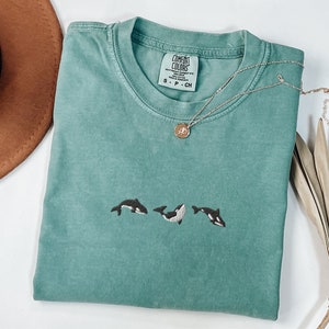 Könnte beinhalten: Ein blaugrünes T-Shirt mit einem schwarzen und weißen gestickten Design von drei Orcas. Das Shirt hat ein Etikett mit der Aufschrift "COMFORT COLORS" und "S - P - CH".