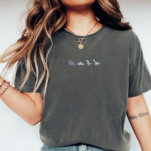 Może przedstawiać: Szary t-shirt z pięcioma małymi, kolorowymi ilustracjami dinozaurów na piersi. Koszulkę nosi osoba z długimi, falującymi brązowymi włosami i złotym naszyjnikiem.