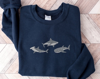 May include: A navy blue sweatshirt with an embroidered design of three sharks. The sharks are a hammerhead, a great white, and a whale shark.